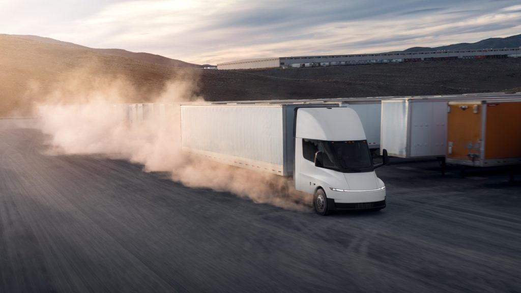 A white electric semi-truck drives on a dusty road past other parked trailers, with dust clouds rising behind it and hills in the background under a cloudy sky.