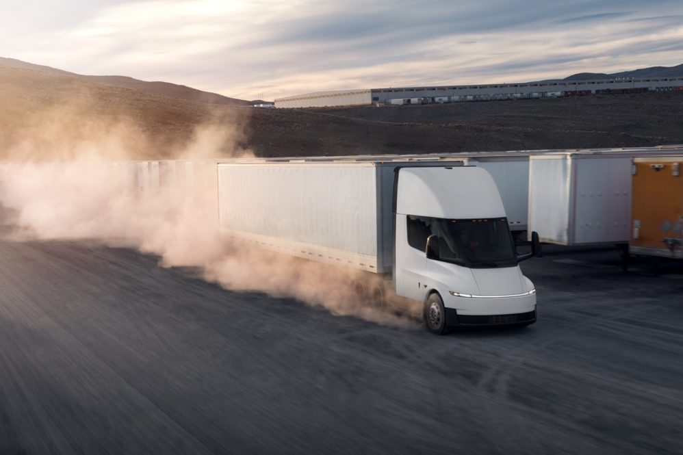 A white electric semi-truck drives on a dusty road past other parked trailers, with dust clouds rising behind it and hills in the background under a cloudy sky.
