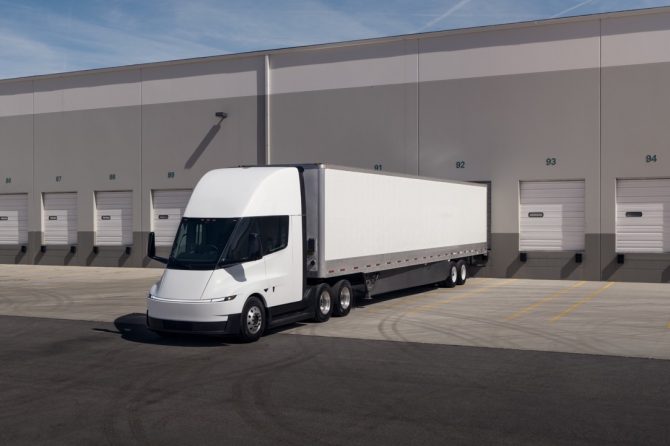 A modern white semi-truck with a long trailer is parked in front of loading docks at a large industrial warehouse. The area is empty and the truck is facing slightly to the left.