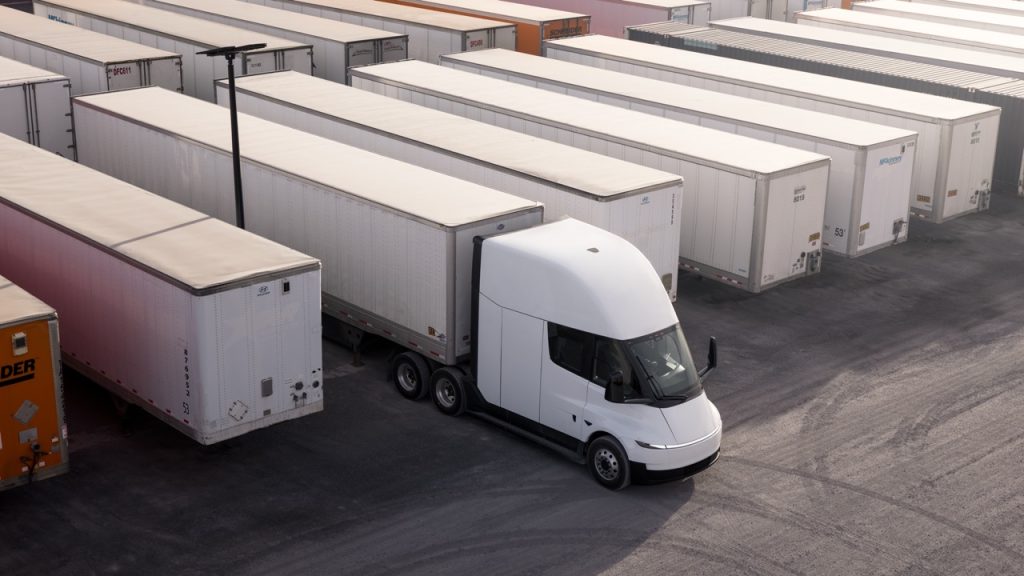 A white semi-truck with a modern design is parked among rows of white freight trailers in a large lot, viewed from above. The trailers are lined up neatly side by side.