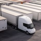 A white semi-truck with a modern design is parked among rows of white freight trailers in a large lot, viewed from above. The trailers are lined up neatly side by side.