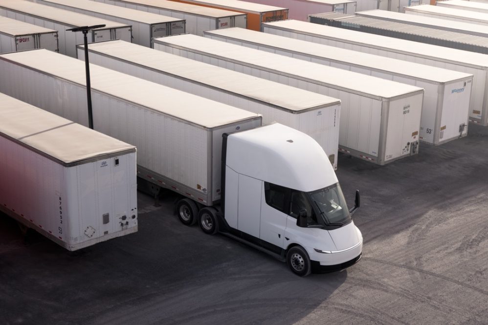 A white semi-truck with a modern design is parked among rows of white freight trailers in a large lot, viewed from above. The trailers are lined up neatly side by side.