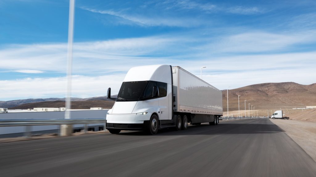 A sleek, modern white semi-truck drives on a paved road through a desert landscape with mountains in the background and a partly cloudy blue sky above.