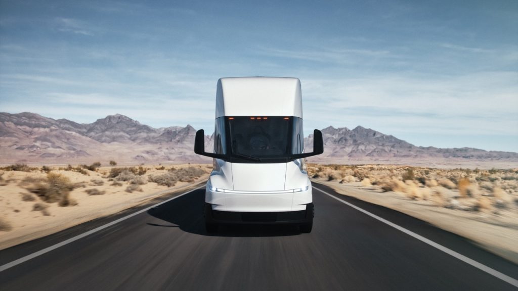 A white semi-truck drives straight down an empty desert highway, with mountains and blue sky in the background. The scene is bright and clear, emphasizing the trucks modern design.