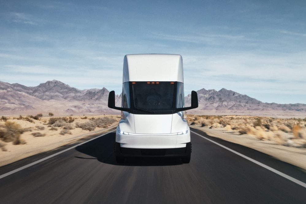 A white semi-truck drives straight down an empty desert highway, with mountains and blue sky in the background. The scene is bright and clear, emphasizing the trucks modern design.