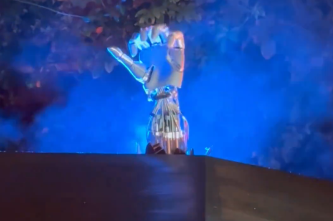 A shiny, metallic robotic hand is raised above a dark surface, illuminated by blue light, with trees and foggy blue smoke in the background at night.