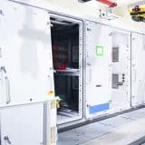 Worker in a hard hat inspecting an open Tesla Megapack 3 beside robotic arms at Megafactory Texas