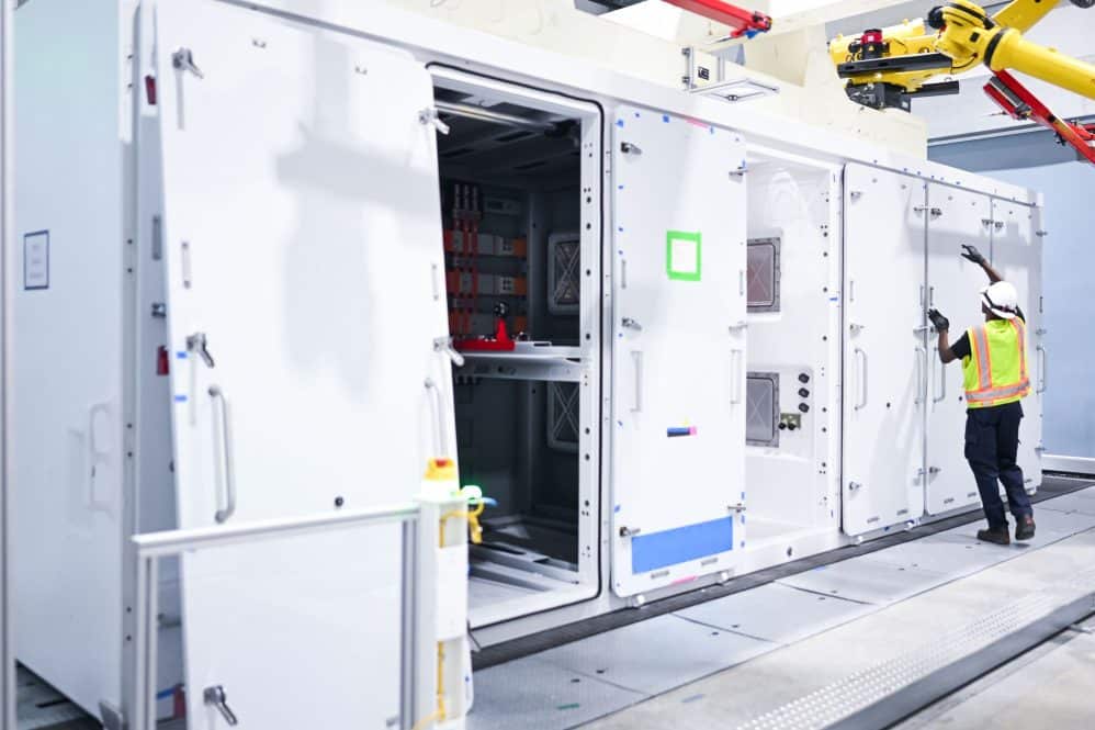 Worker in a hard hat inspecting an open Tesla Megapack 3 beside robotic arms at Megafactory Texas