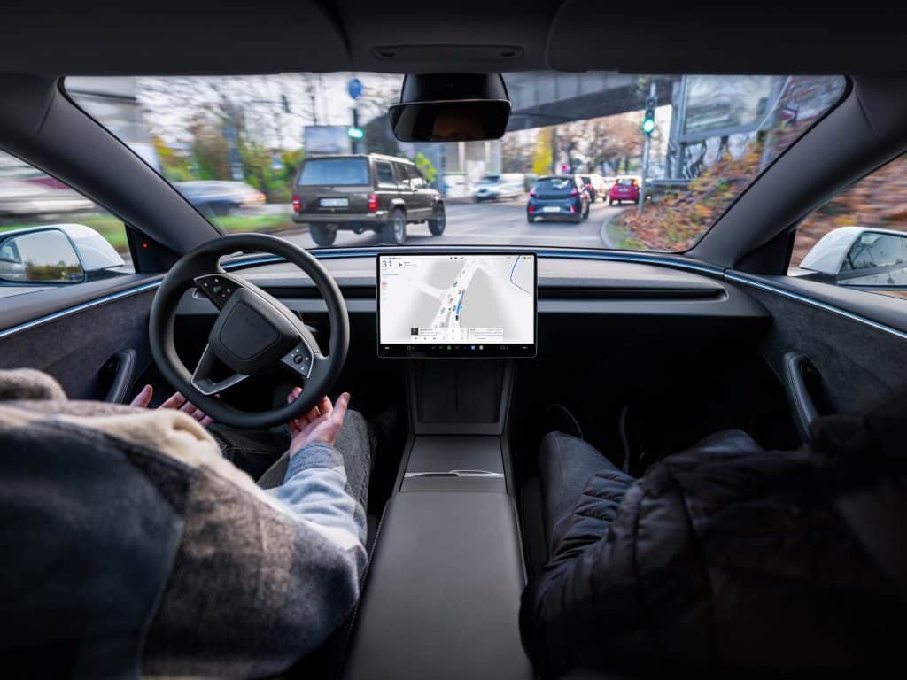 A driver's hands rest off the steering wheel of a Tesla as FSD (Supervised) steers through city traffic, with the navigation map on the centre touchscreen