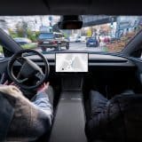 A driver's hands rest off the steering wheel of a Tesla as FSD (Supervised) steers through city traffic, with the navigation map on the centre touchscreen