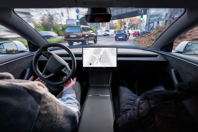 A driver's hands rest off the steering wheel of a Tesla as FSD (Supervised) steers through city traffic, with the navigation map on the centre touchscreen