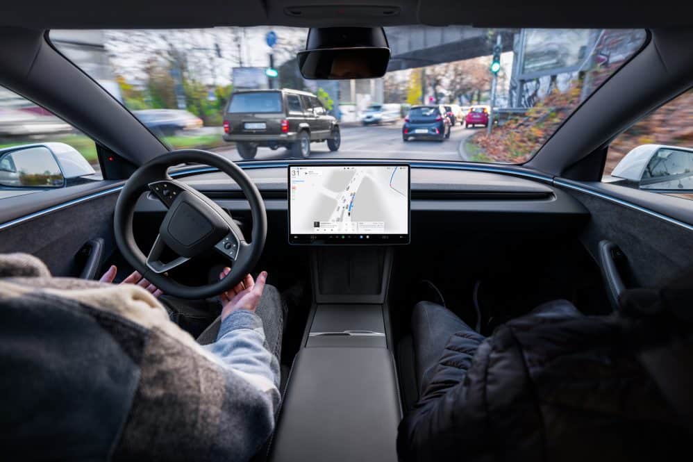 A driver's hands rest off the steering wheel of a Tesla as FSD (Supervised) steers through city traffic, with the navigation map on the centre touchscreen