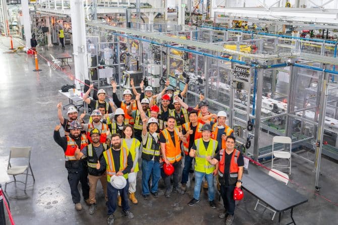 Tesla employees in hard hats and safety vests cheer in front of the first-generation Optimus humanoid robot production line in Fremont, California
