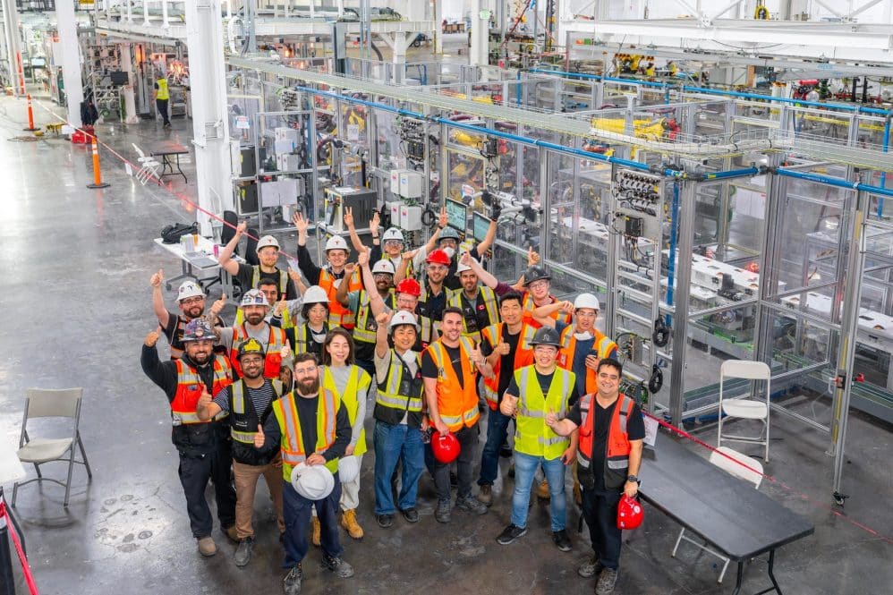 Tesla employees in hard hats and safety vests cheer in front of the first-generation Optimus humanoid robot production line in Fremont, California